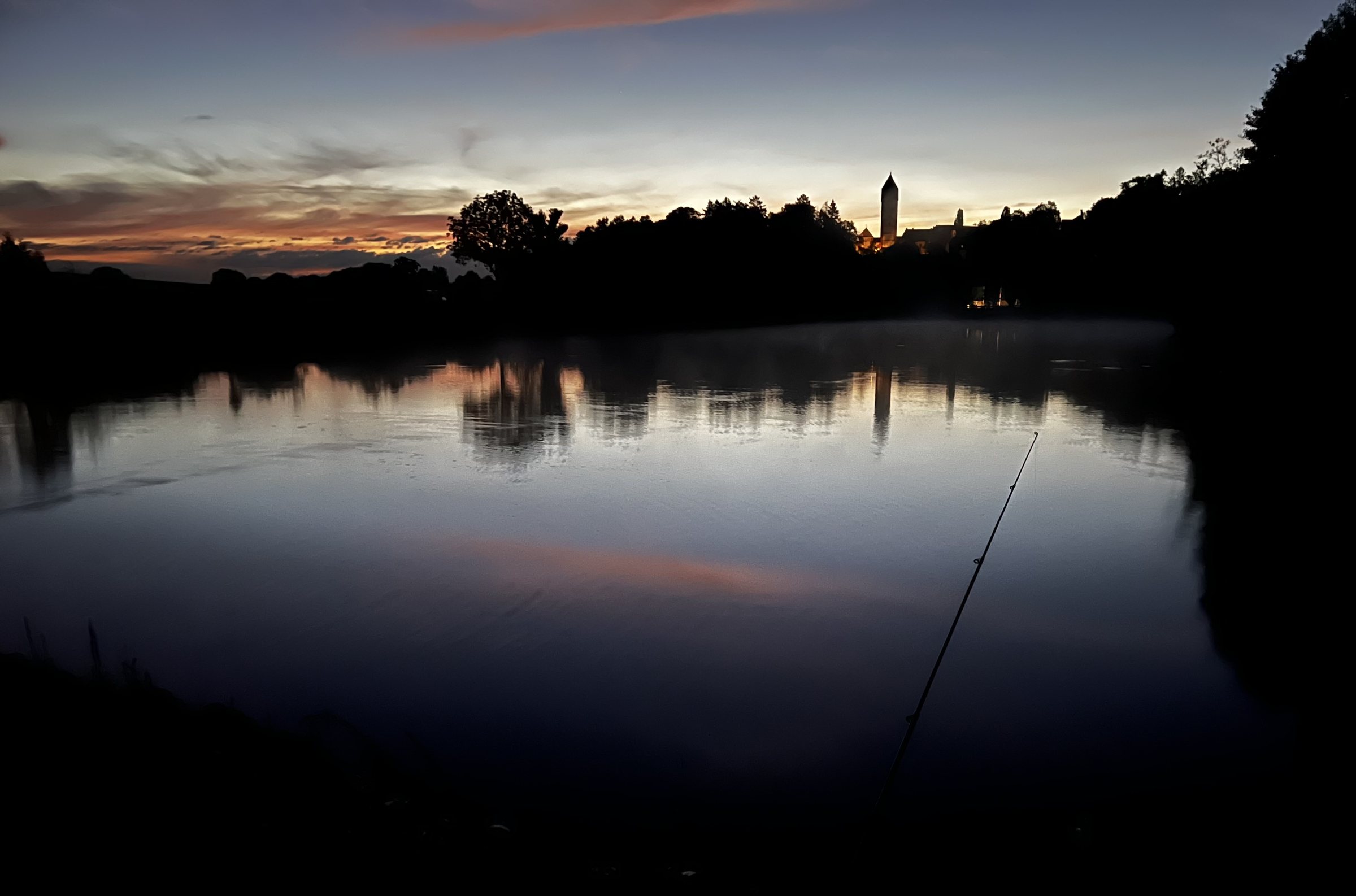 Angelrute am Ufer eines Weihers in der Morgendämmerung, der rot gefärbte Himmel spiegelt sich im Wasser