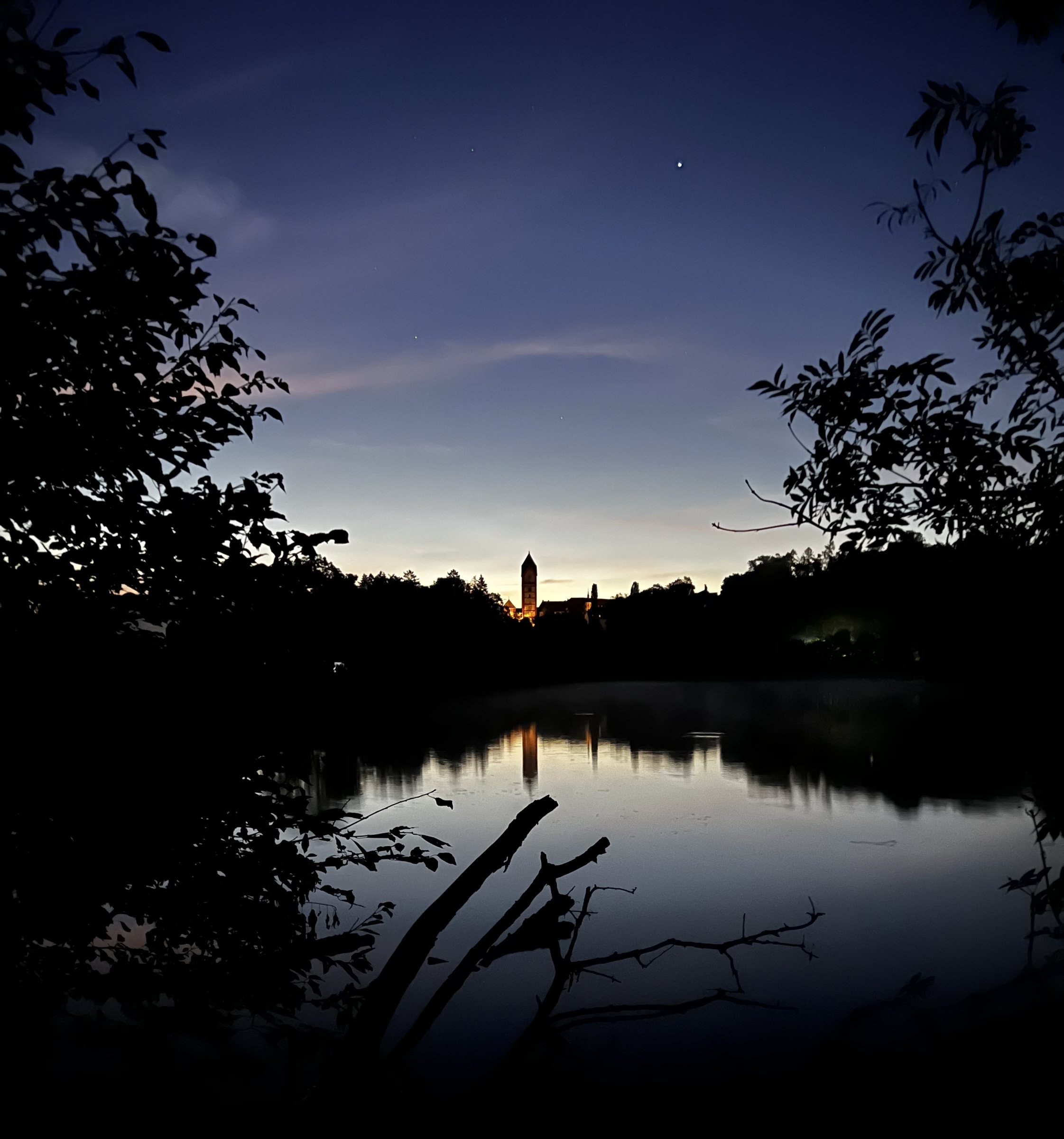 Blick durch Zweige über einen Weiher in der Morgendämmerung, am Horizont ein beleuchteter Kirchturm